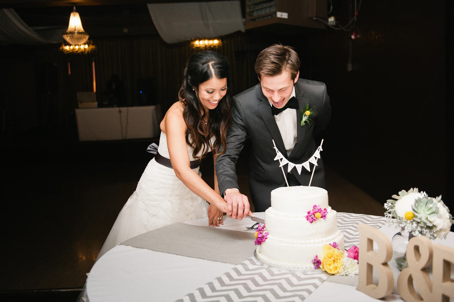 A bride and groom cut a white tiered wedding cake decorated with flowers and bunting, showcasing one of the latest wedding cake styles, at a reception table with a gray chevron runner and floral arrangement.