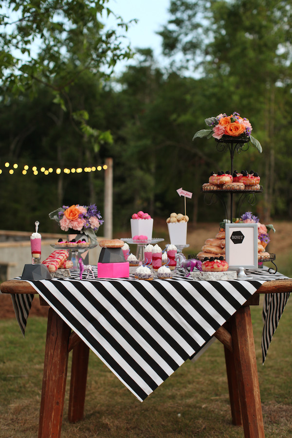 Outdoor dessert table with a black and white striped tablecloth, assorted pastries, flowers, string lights, and an array of wedding cake styles showcased in the background.
