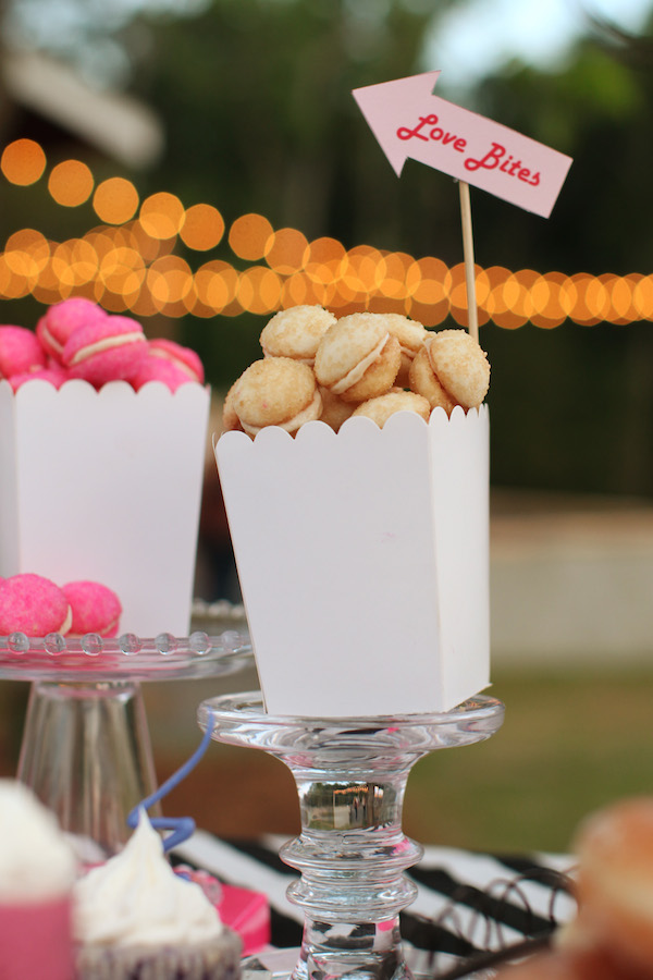 A white container labeled "Love Bites" holds small round cookies, displayed on a glass stand alongside other pink and white desserts, perfectly complementing elegant wedding cake styles in the background.