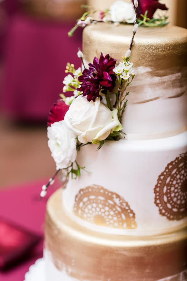 Three-tier white and gold cake decorated with maroon and white flowers and intricate gold patterns, showcasing elegant wedding cake styles.
