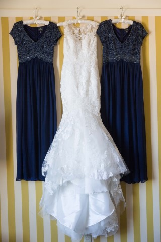 Lace mermaid wedding dress hanging between two navy bridesmaid dresses on striped wall