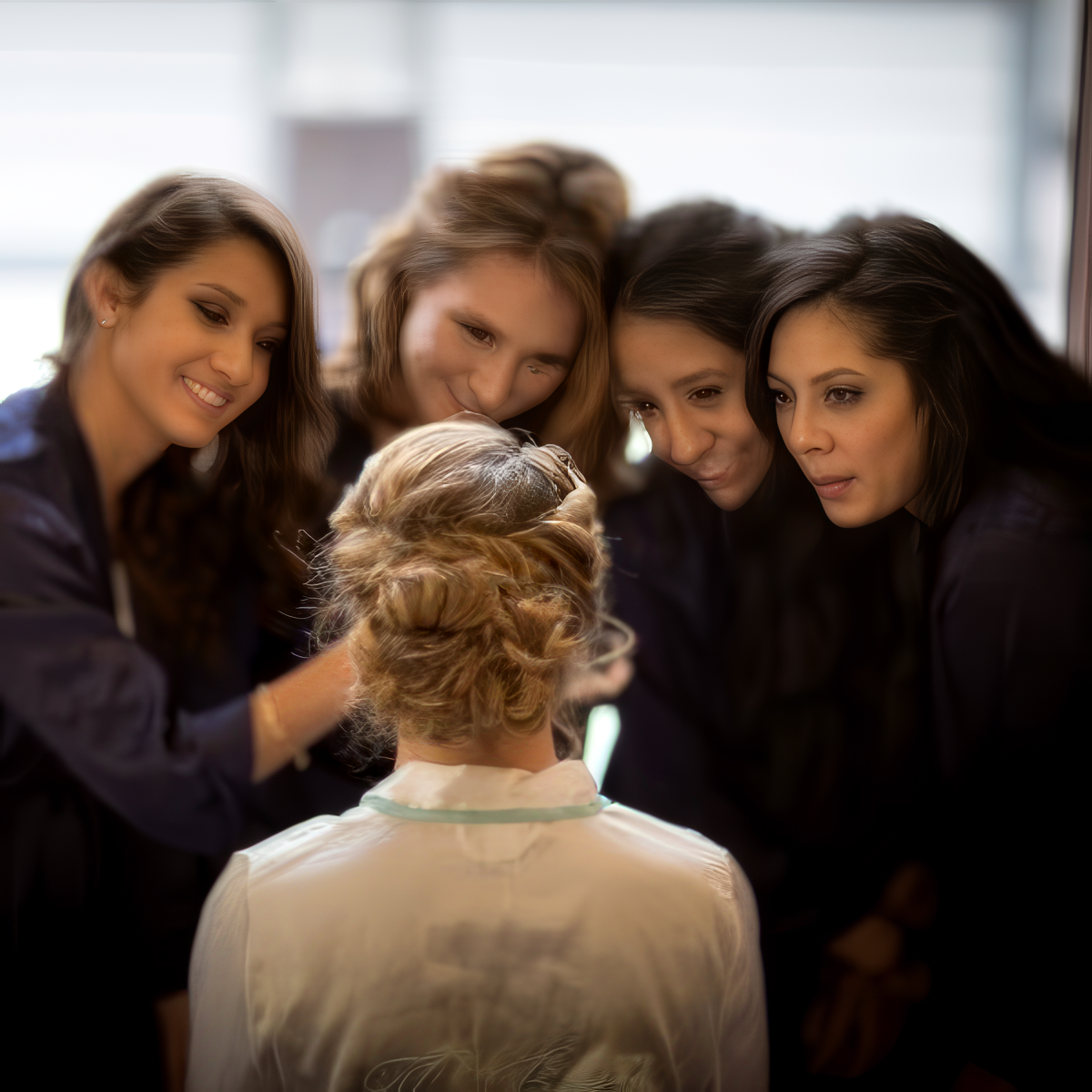 Four women stand closely together, smiling and looking at another woman with styled hair who is seated with her back to the camera during a wedding at Oakhurst Country Club.