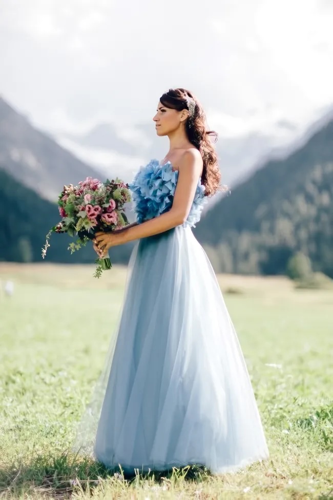 A woman in a light blue gown holds a bouquet of flowers while standing on a grassy field, mountains rising majestically behind her during this Mont Blanc styled shoot.