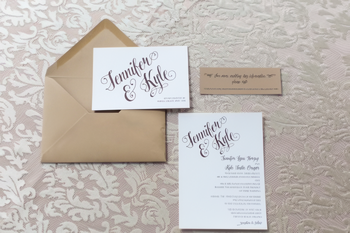 A beige envelope with three wedding invitation cards displaying "Jennifer & Kyle" in cursive rests on a patterned tablecloth, setting the scene for a wedding with custom wood hangers.