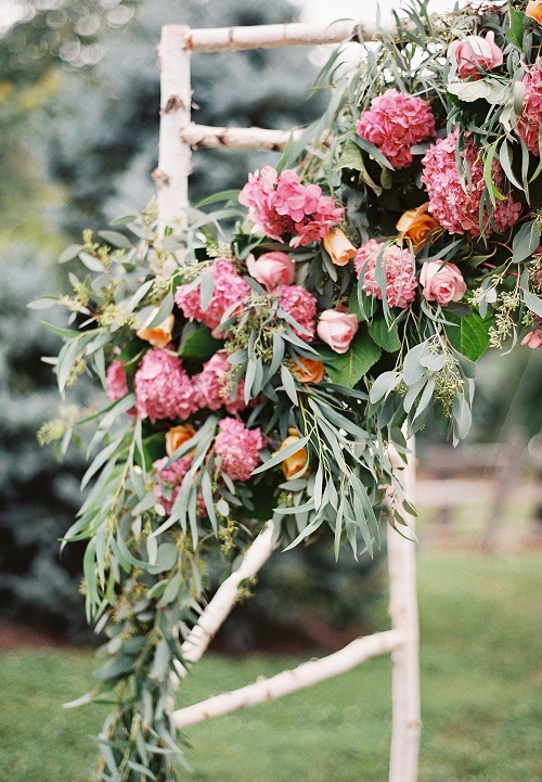 Rustic wedding ceremony arch made of white birch branches decorated with pink hydrangeas, peach roses, and cascading eucalyptus greenery