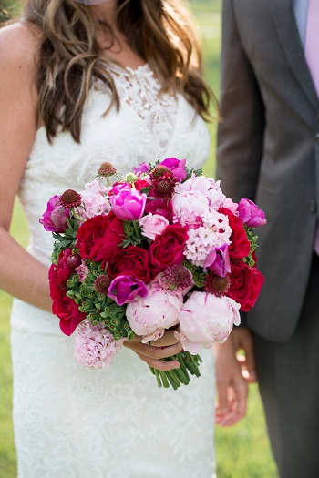 Bride in lace gown holding garden-style bouquet with deep red roses, hot pink peonies, blush peonies, burgundy scabiosa, and green hypericum berries