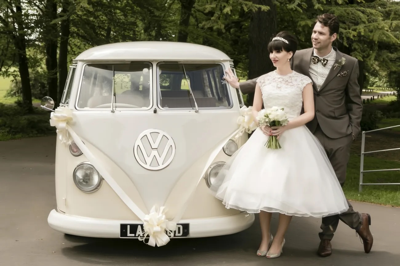 A bride and groom stand beside a vintage 1960s VW van decorated with ribbons, surrounded by greenery, perfect for a stylish theme wedding.