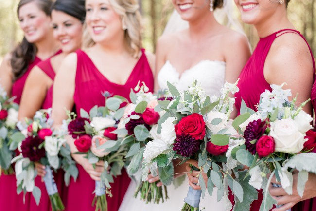 Bridesmaids in bright red-pink dresses and bride in sweetheart gown holding bouquets of red roses, white roses, burgundy dahlias, and silvery eucalyptus
