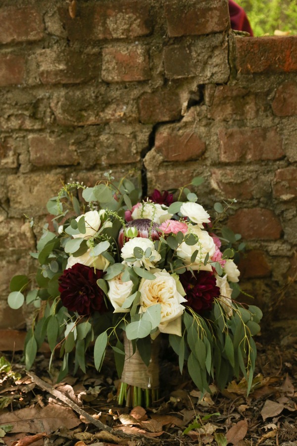 Standalone bridal bouquet against rustic brick wall featuring burgundy dahlias, cream and yellow roses, pink protea, and cascading eucalyptus