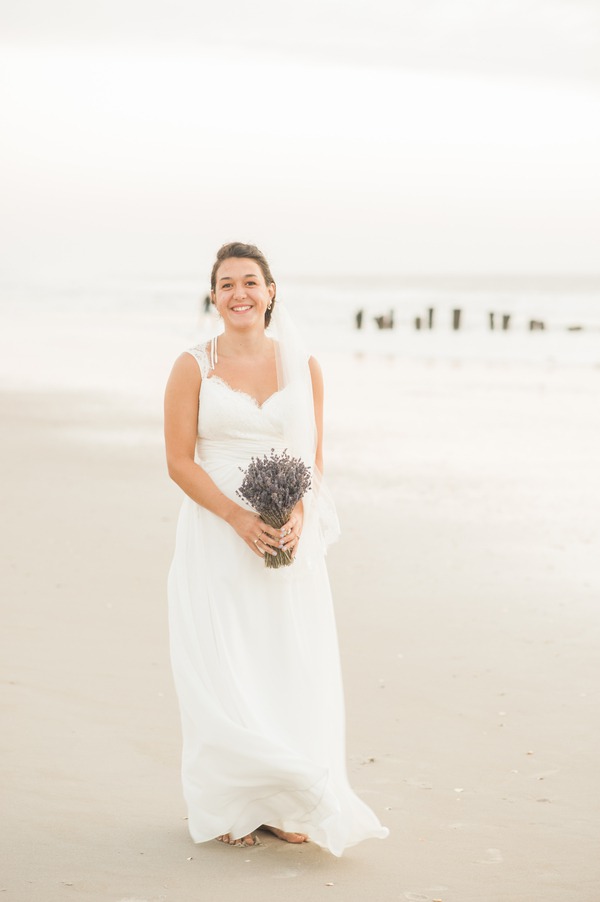 Smiling bride standing barefoot on beach sand holding dried lavender bouquet tied with twine