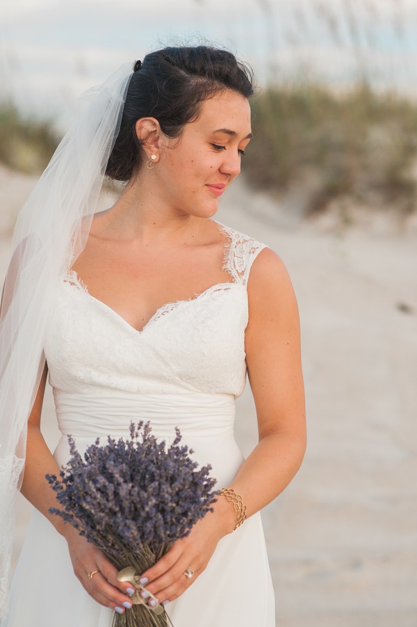 Bride in lace cap sleeve gown looking down at dried lavender bouquet on beach with dunes in background