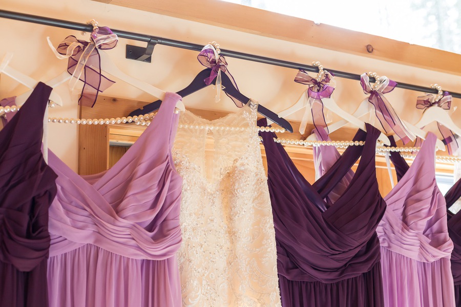 Five bridesmaid dresses in alternating soft lilac and deep plum shades hanging on a rack with purple ribbon bows and pearl strands, with the bride's gown in the center