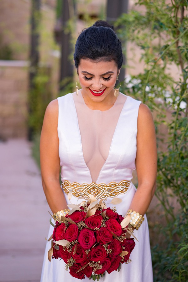 Bride in deep V-neck gown with gold beaded belt holding compact red rose bouquet with gold-painted leaves and seeded eucalyptus