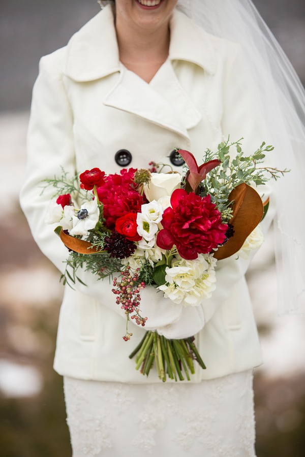 Bride in white peacoat and veil holding loose winter bouquet with red peonies, white anemones, ranunculus, magnolia leaves, and pink pepperberries