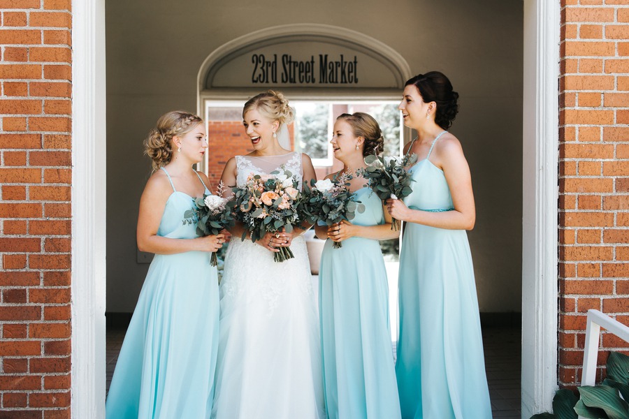 Three bridesmaids in matching ice blue floor-length spaghetti-strap chiffon gowns standing with the bride in a red brick archway holding blush and eucalyptus bouquets