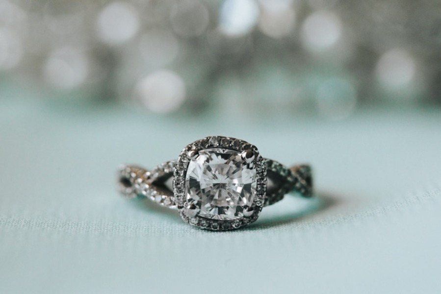 Cushion-cut diamond in a dark metal twisted pavé band with two-tone black and white diamond halo on a teal background with bokeh