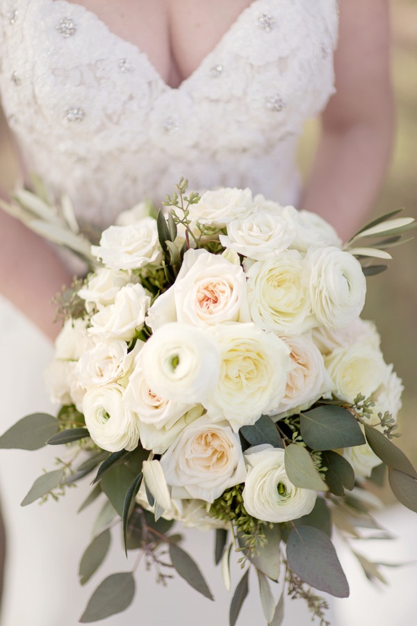 Bride holding classic all-white bouquet with garden roses, ranunculus, and olive leaf greenery against beaded sweetheart neckline gown