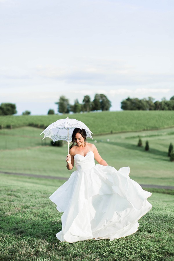 Bride holding white lace parasol wearing sleeveless white satin ball gown with sweetheart neckline running through vineyard