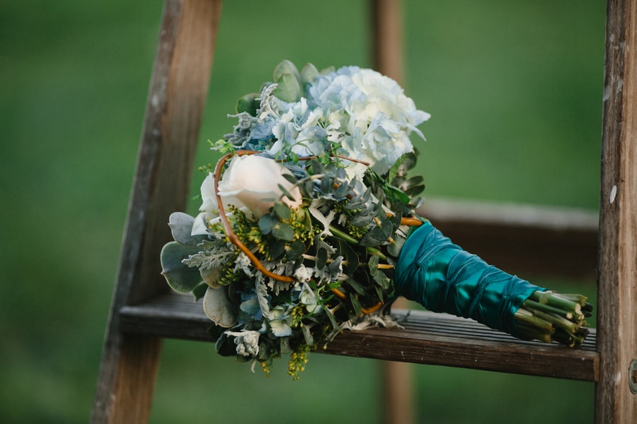 Blue hydrangea bouquet with dusty miller, craspedia, eucalyptus, and curly willow wrapped in teal satin ribbon on rustic ladder