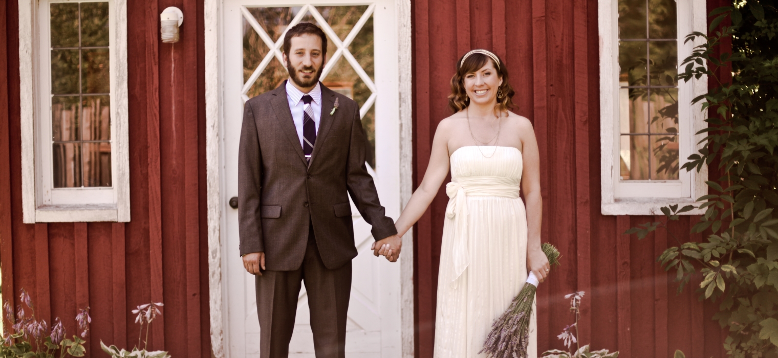 Bride and groom holding hands in front of red barn, bride carrying fresh lavender bouquet