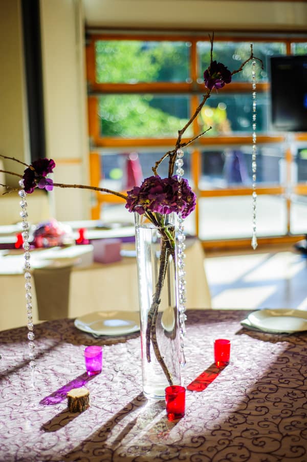 Tall centerpiece with deep purple hydrangeas and bare branches with hanging crystals on purple tablecloth