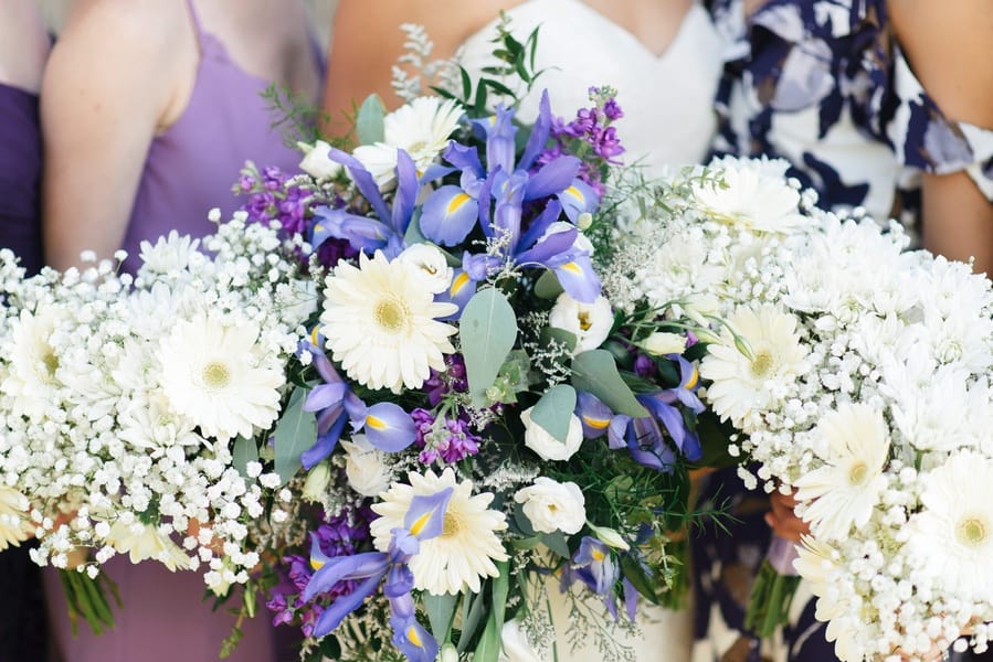 Bridal bouquet with purple iris, white gerbera daisies, purple stock, baby's breath, and eucalyptus with bridesmaids holding simpler baby's breath bouquets