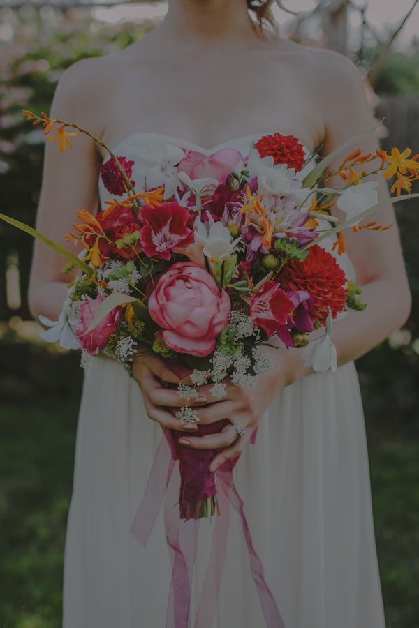 Bride in sweetheart neckline gown holding loose garden bouquet with pink peonies, red dahlias, orange crocosmia, white cosmos, Queen Anne's lace, and trailing burgundy ribbon