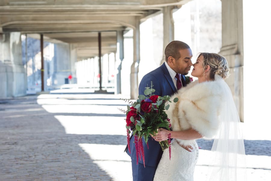Bride in an ivory fur stole with a cascading red bouquet leaning toward her groom under an urban overpass