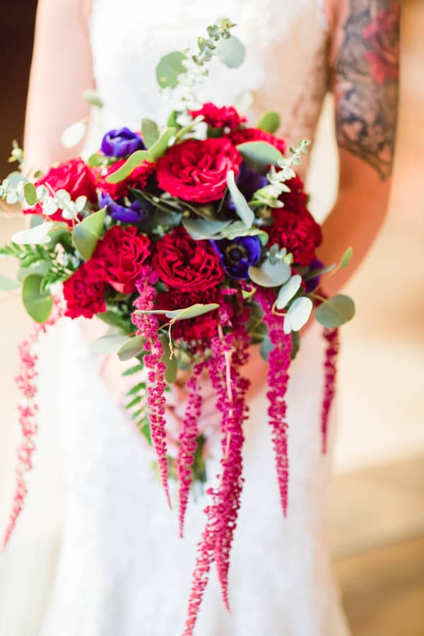 Tattooed bride holding cascading bouquet with red garden roses, burgundy carnations, purple anemones, and dramatic trailing magenta amaranthus