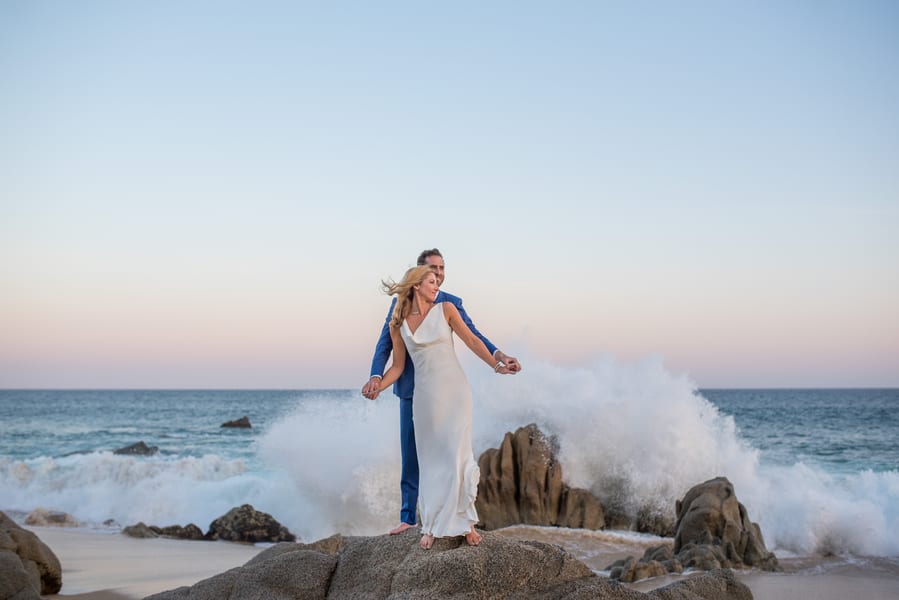 Couple balanced on rocks with wave crashing, bride in minimalist slip dress