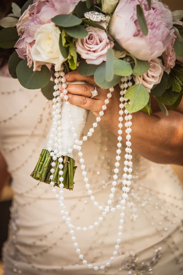 Close-up of bride holding lavender and blush peony bouquet with mauve roses and eucalyptus, draped with long pearl strands against beaded gown