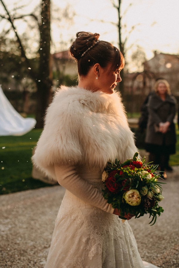 Bride with elegant updo wearing fluffy white fur stole holding red and cream bouquet in snowy Italian setting