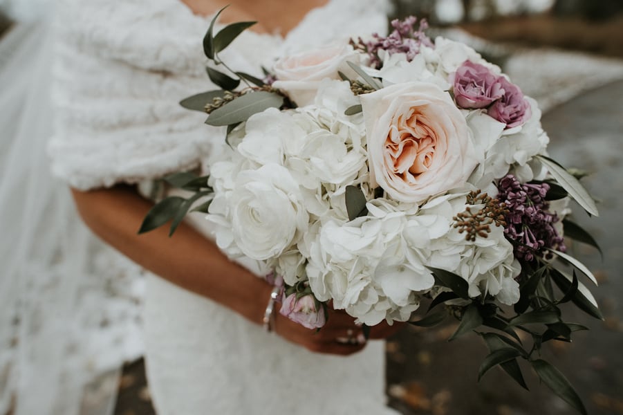 Bride in white fur stole holding bouquet of white hydrangeas, blush garden roses, and mauve lilac accents