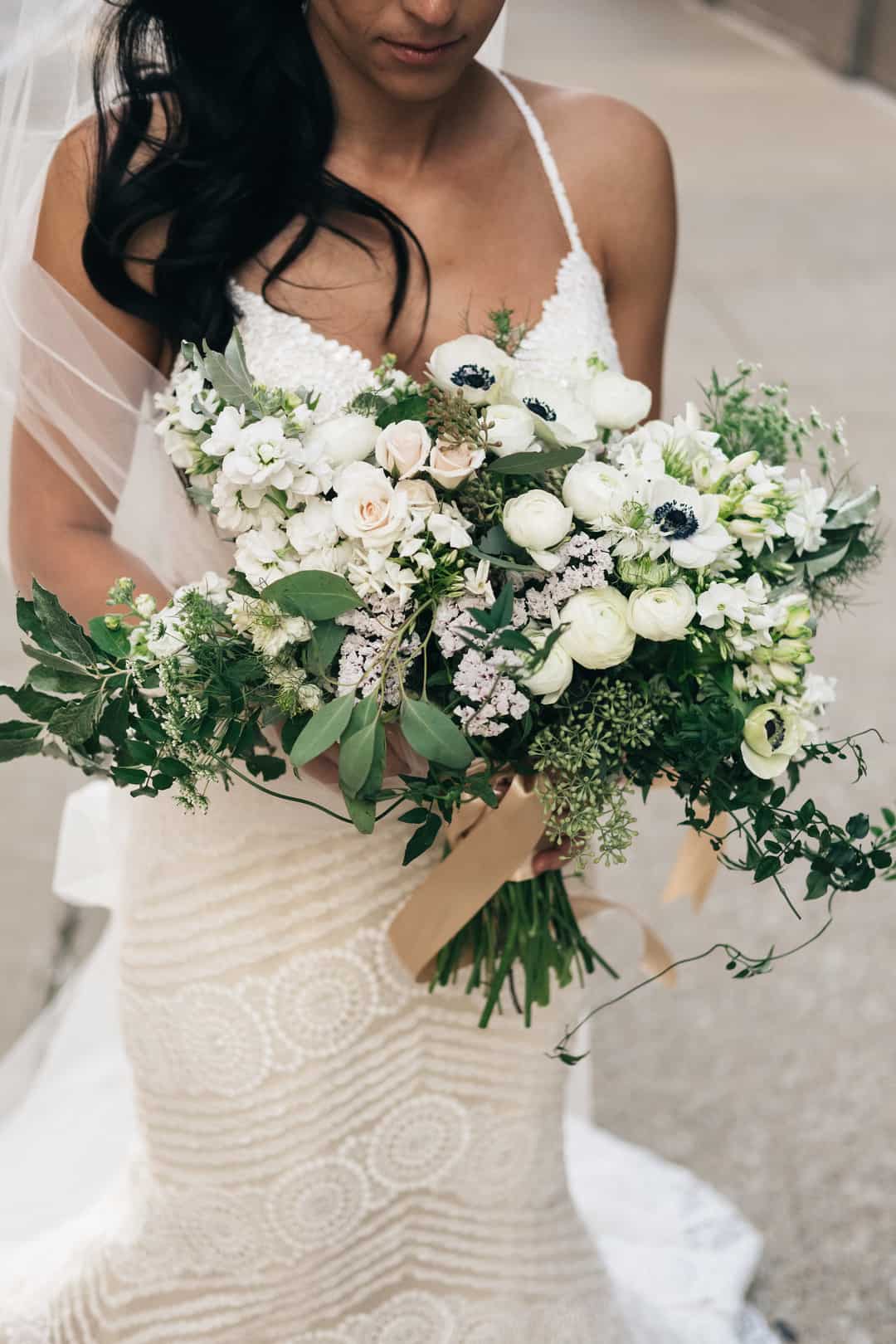 Bride in textured halter gown holding large cascading bouquet of white roses, ranunculus, anemones, stock, and trailing greenery with tan ribbon