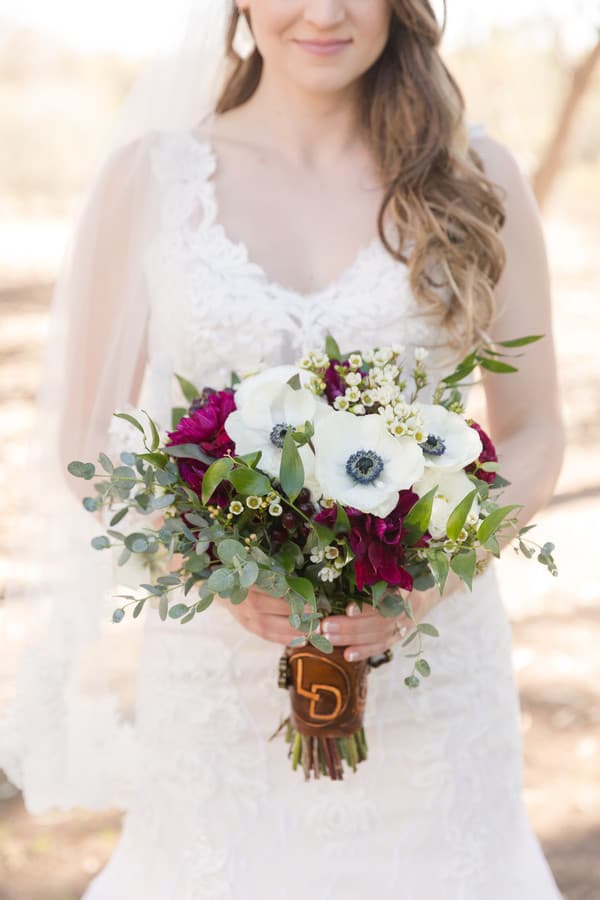 Bride in lace V-neck gown holding bouquet with white anemones, burgundy dahlias, waxflower, and eucalyptus wrapped with brown leather and monogram charm