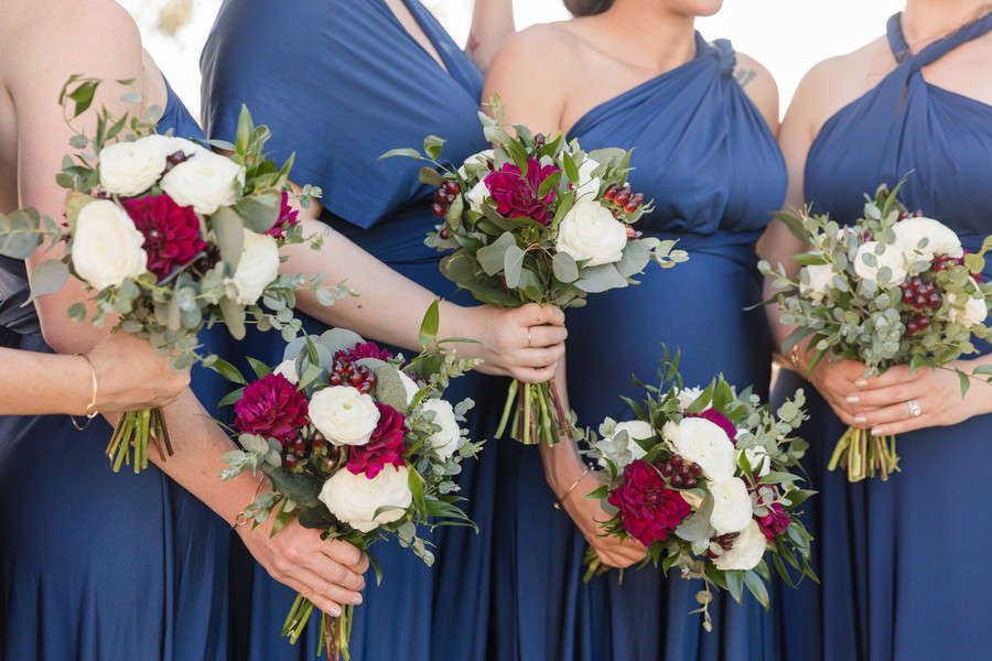 Bridesmaids in navy blue convertible wrap dresses holding coordinating bouquets with white ranunculus, magenta dahlias, and eucalyptus