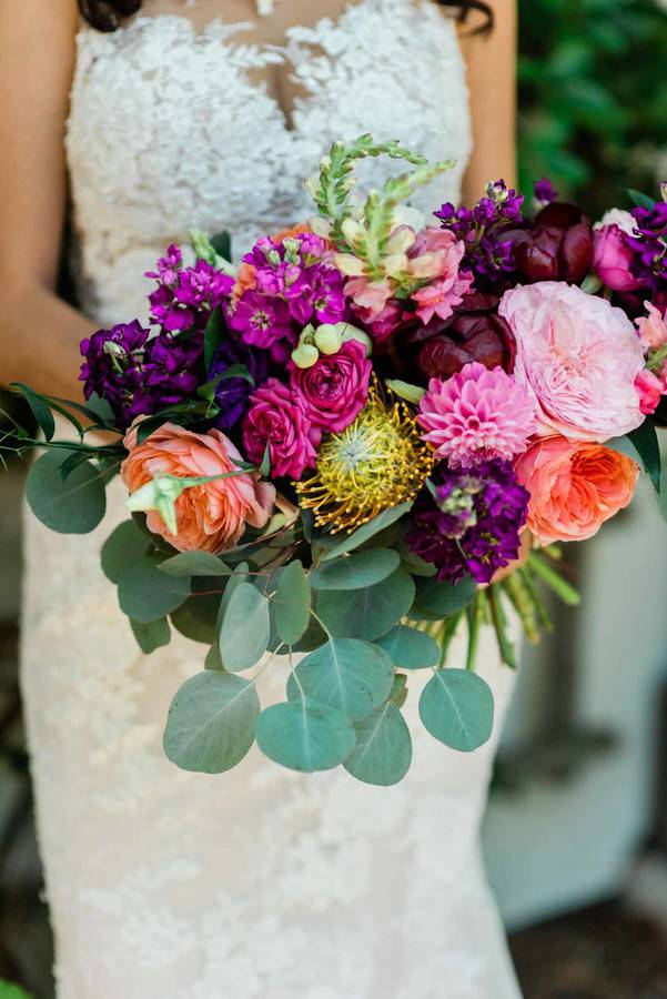 Bride in lace gown holding colorful bouquet with purple stock, hot pink roses, coral garden roses, pink dahlia, yellow pincushion protea, and silver dollar eucalyptus