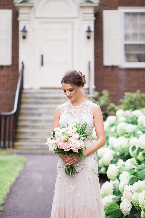 Bride in vintage-inspired beaded high-neck blush gown holding pink and white rose bouquet in front of historic brick building with white hydrangeas