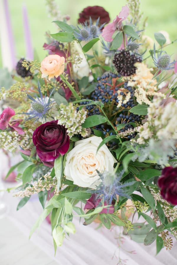 Lush organic floral arrangement with cream roses, burgundy ranunculus, peach spray roses, blue thistle, privet berries, astilbe, and mixed greenery