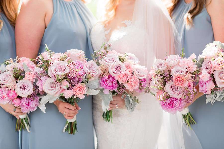 Bride and bridesmaids in dusty blue dresses holding coordinating pink bouquets with roses, carnations, peonies, and waxflower