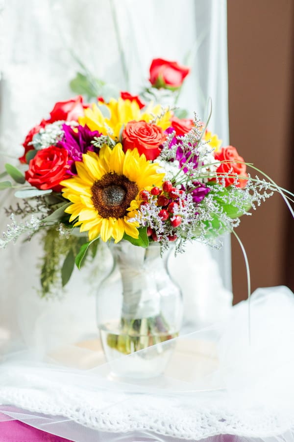 Vibrant wedding bouquet with sunflowers, red roses, coral spray roses, and magenta blooms in a vase