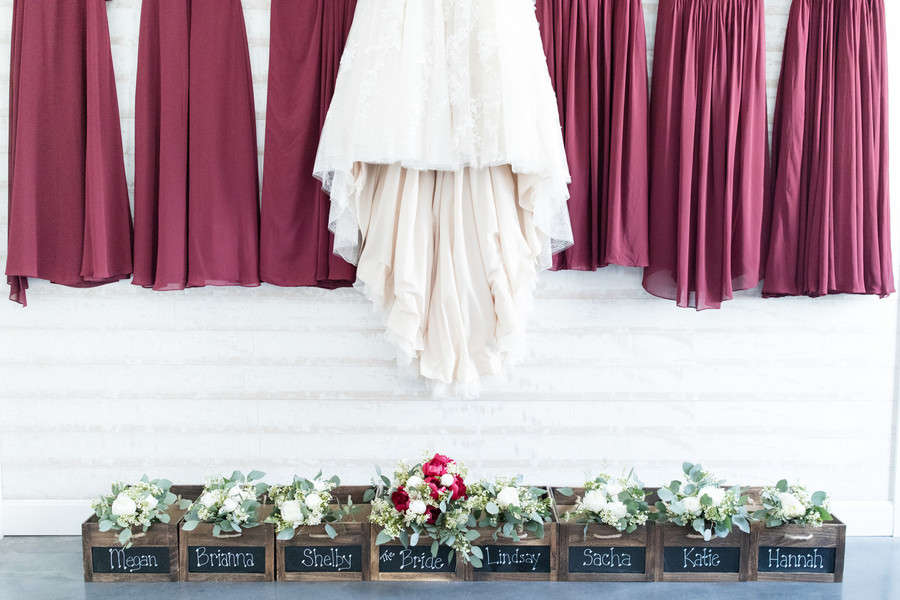 Bridal party bouquets displayed in wooden boxes with chalkboard name labels, burgundy bridesmaid dresses hanging above, bride's bouquet with red roses in center