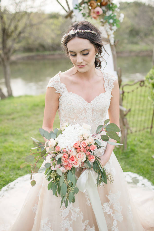 Bride in champagne lace ball gown with crystal headpiece holding cascading bouquet of coral spray roses, white hydrangea, and eucalyptus by the lake