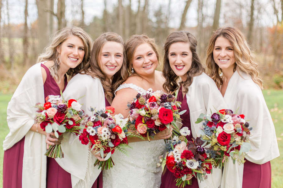 Bride and four bridesmaids in burgundy dresses with cream shawls holding colorful bouquets with red roses, coral blooms, blue thistle, white anemones, and mixed greenery