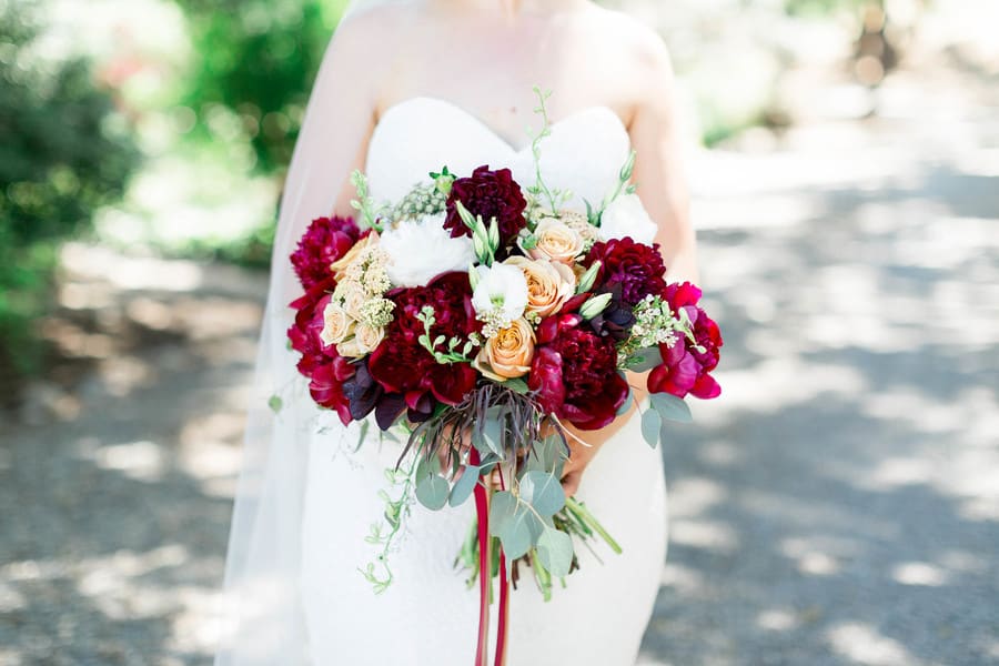 Close-up of bridal bouquet with deep burgundy peonies, peach roses, white ranunculus, eucalyptus, and trailing burgundy ribbon