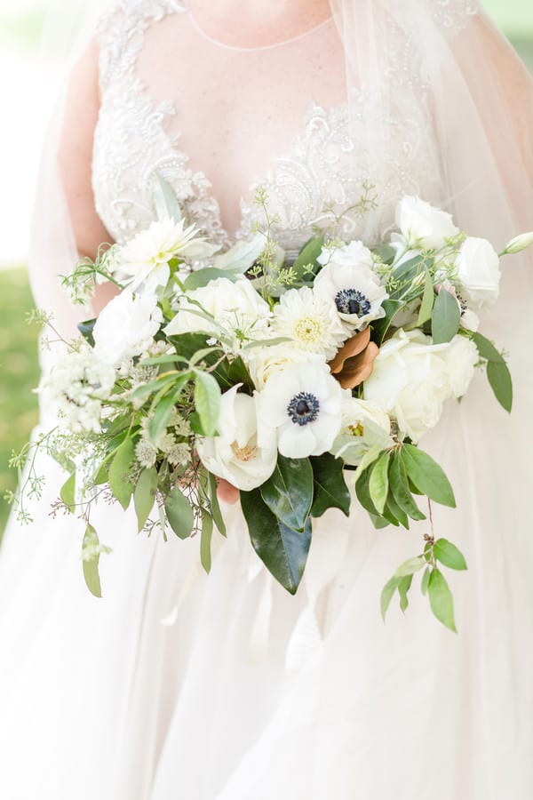 Bride in illusion lace bodice gown holding loose organic bouquet of white roses, anemones, dahlias, and mixed greenery