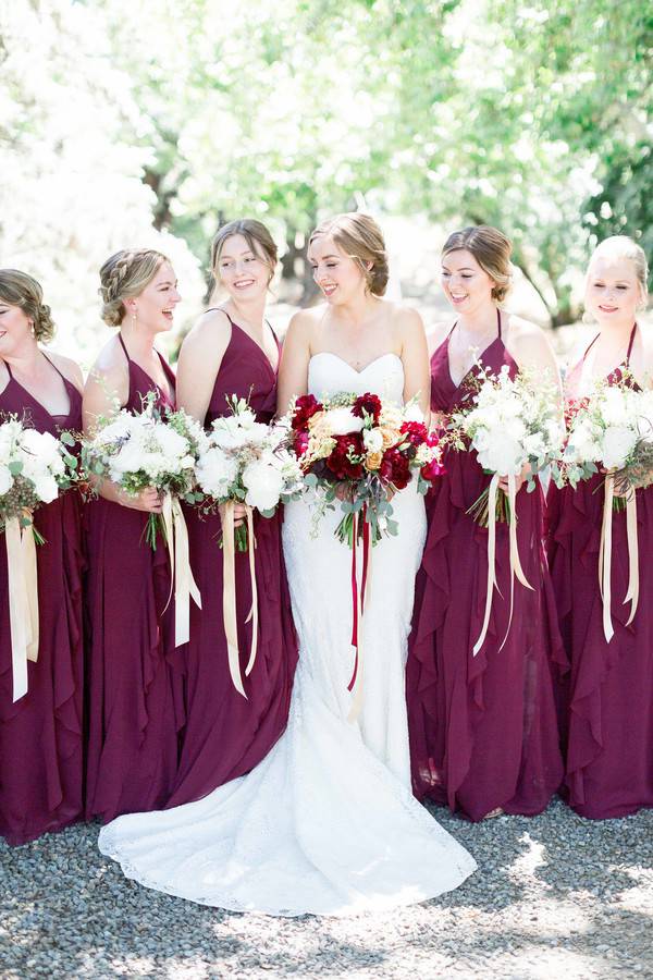 Bride in sweetheart lace gown surrounded by bridesmaids in burgundy halter dresses, bride holding burgundy and gold bouquet, bridesmaids with white and green arrangements tied with champagne ribbon