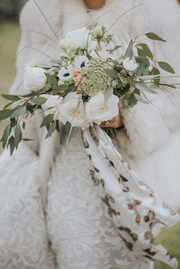 Bride in white fur stole and textured gown holding loose bouquet of white roses, anemones, and queen anne's lace with floral print ribbon