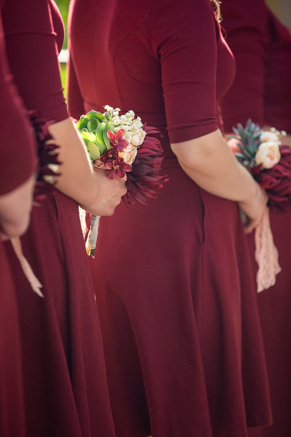 Behind view of bridesmaids in long-sleeve burgundy dresses holding petite posies with succulents, protea, and peach blooms