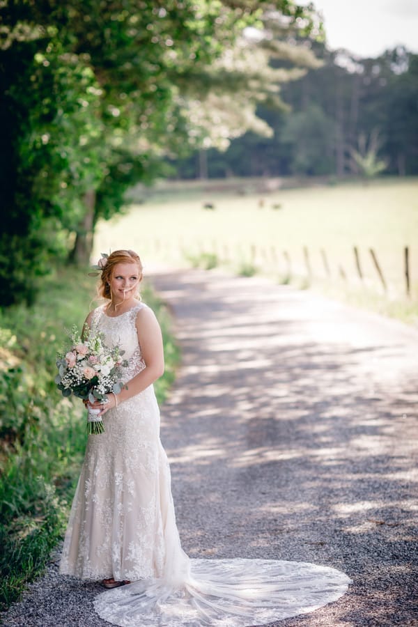 Bride in high-neck lace gown with circular train standing on country road holding soft pink and white bouquet with roses, stock, and eucalyptus